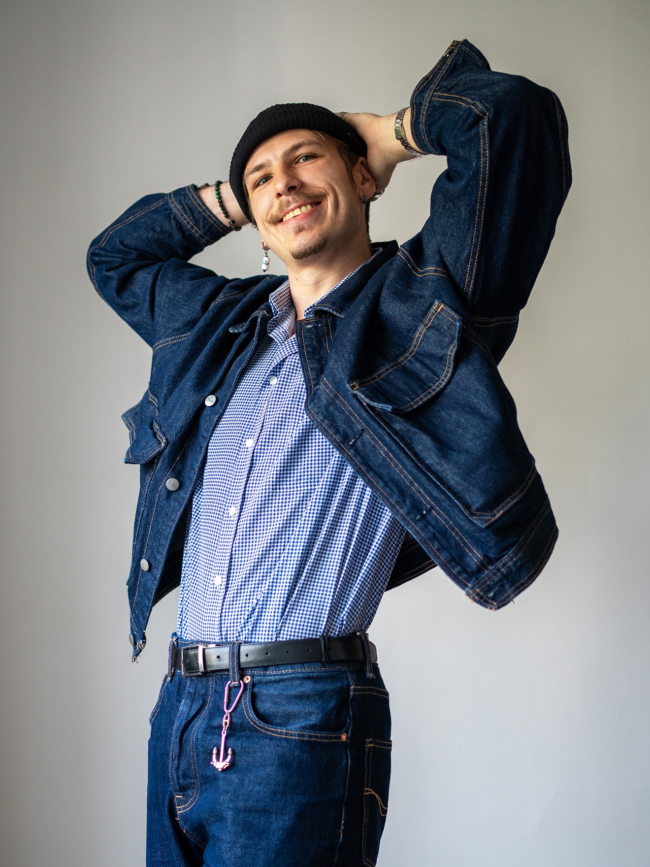Porträt des lächelnden Tanztrainers Marcel in Jeansjacke, Beanie und Ohrring, mit den Händen hinter dem Kopf.